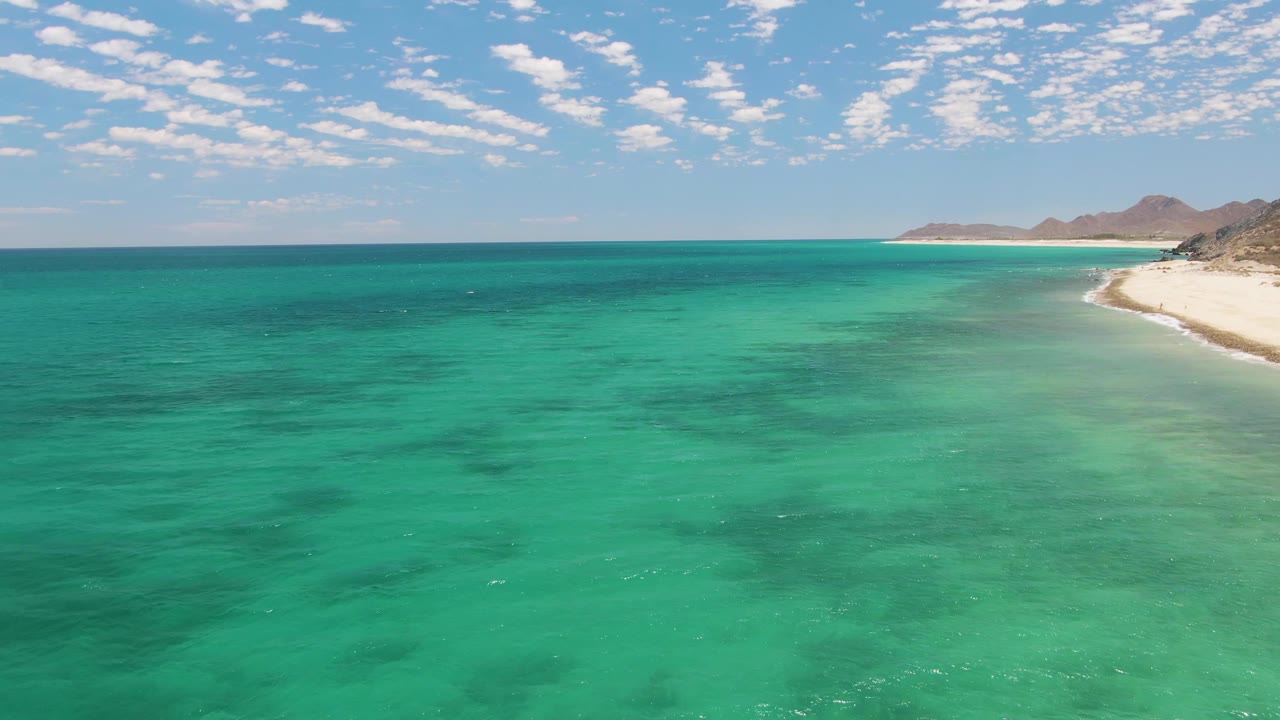 Aerial view at Barakas Beach with open turquoise water across the frame, a small strip of white sand at the upper right, distant hills, broken clouds, and no visible people.