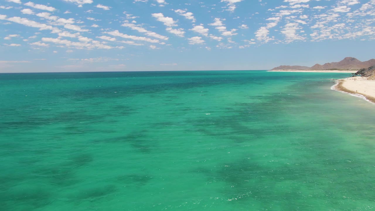 Aerial coastal view from Barakas Beach, Baja California Sur, with calm turquoise water in the foreground, distant white sand and hills at right, scattered clouds, and no visible people.