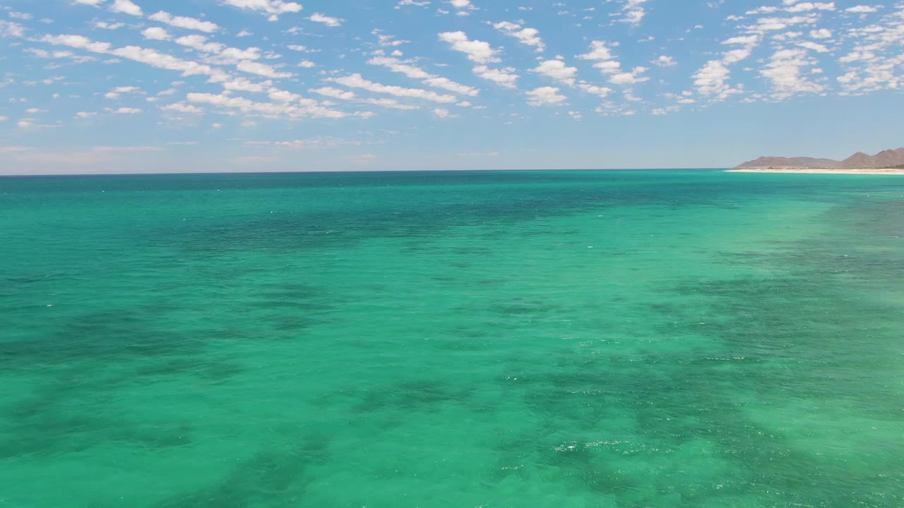 Second frame showing clear turquoise ocean at Barakas Beach, Baja California Sur, with subtle wave texture, darker seafloor areas, a narrow sandy shoreline, low hills, clouds, and no visible people.