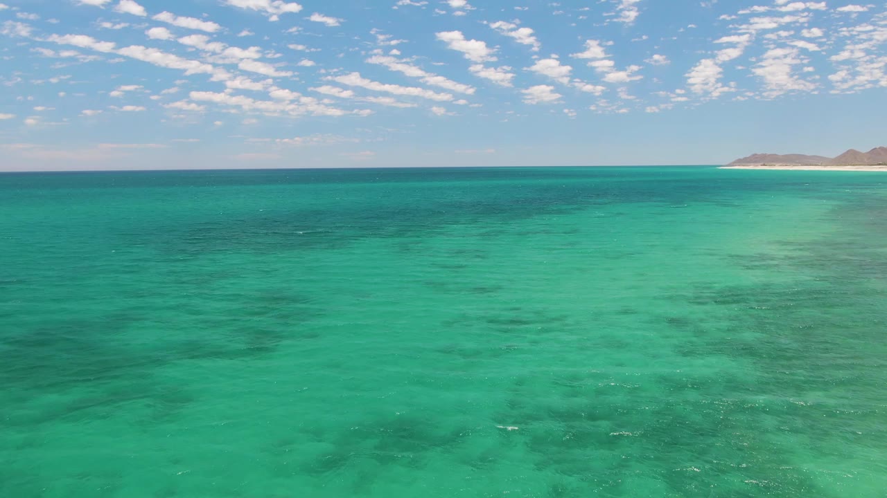 Third frame from Barakas Beach, Baja California Sur, with shallow turquoise sea, pale sand-colored water near the right horizon, darker underwater patches, distant coastline, clouds, and no visible people.