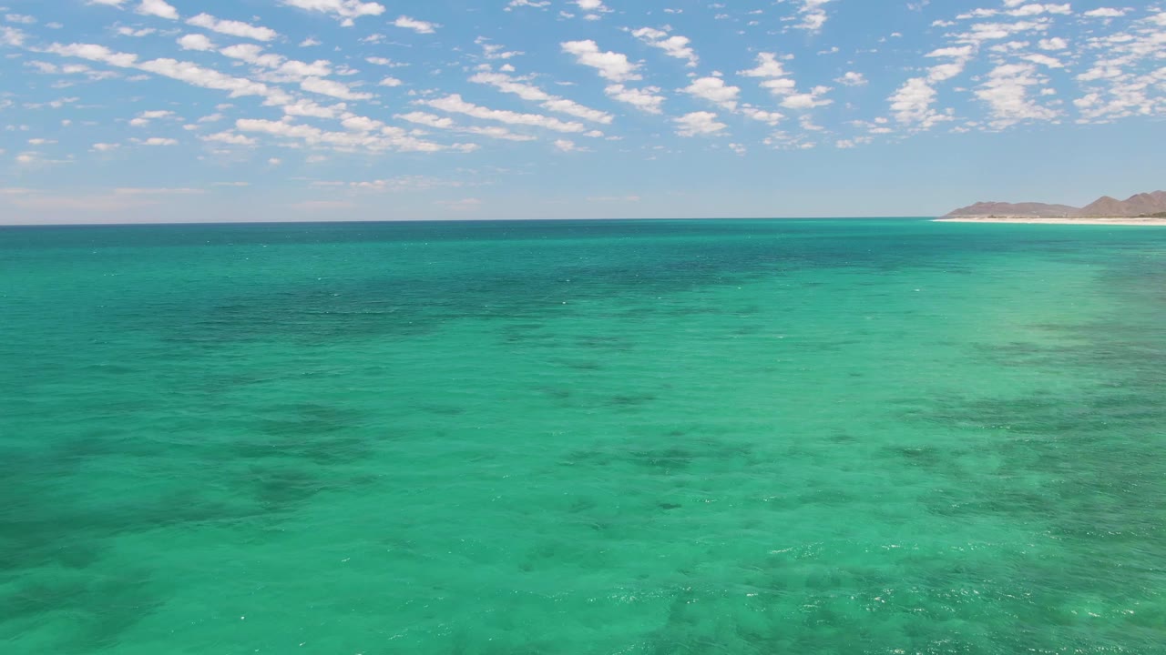 Fourth frame of Barakas Beach, Baja California Sur, with a broad turquoise water field, soft ripples, darker subsurface patches, distant sand and land at the horizon, scattered clouds, and no visible people.