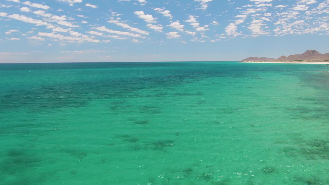 Wide aerial view over turquoise water at Barakas Beach, Baja California Sur, with darker underwater patches, distant brown hills on the right horizon, and no visible people.