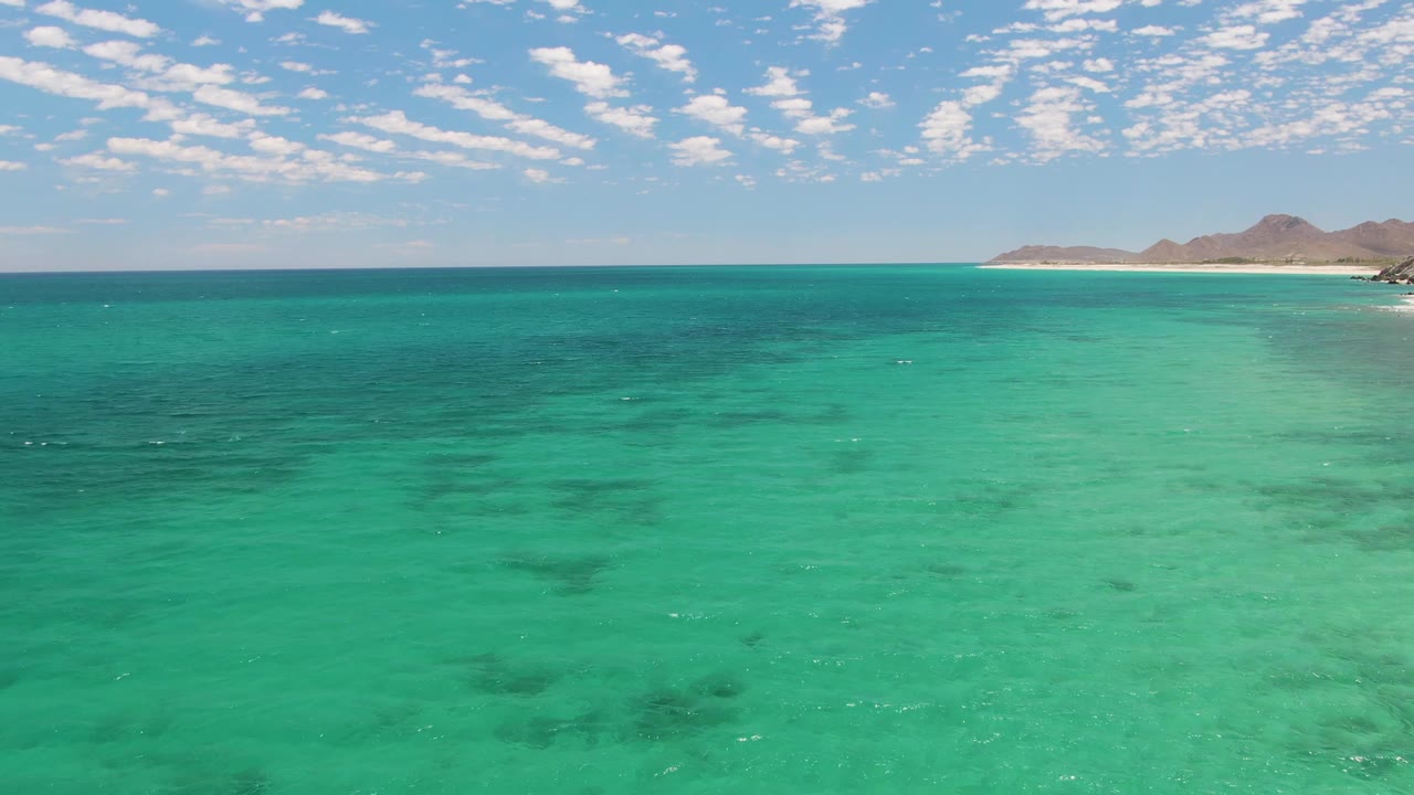 Aerial coastal frame at Barakas Beach with clear turquoise sea in the foreground, a pale sandy strip and rocky brown hills entering from the right, and no visible people.
