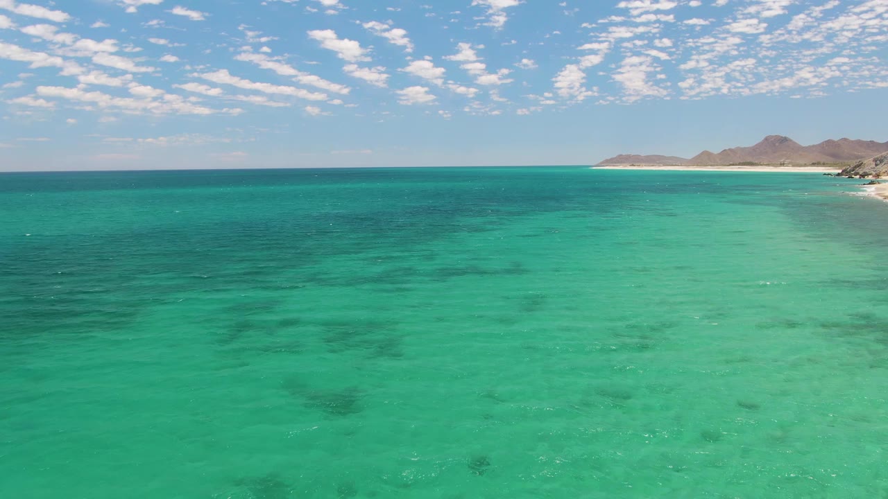 Barakas Beach aerial view showing turquoise water across the foreground, a thin light beach along the right background, low brown hills beyond it, and no visible people.