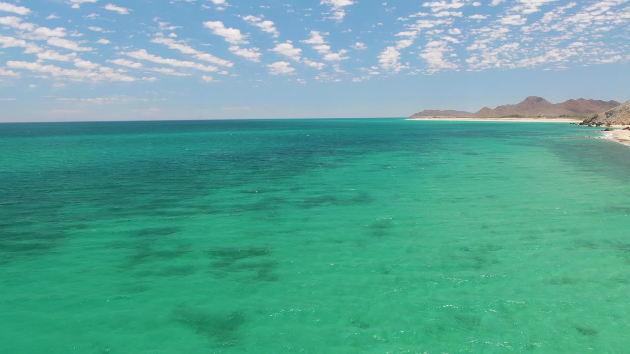 Wide turquoise bay with sandy shore and hills beneath a clouded sky.