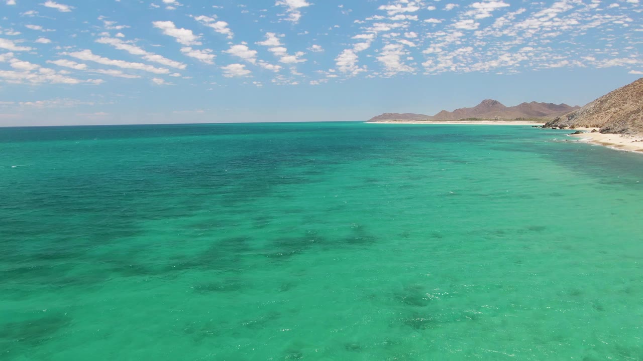 Turquoise ocean beside a sandy shore and rocky hills under scattered clouds.