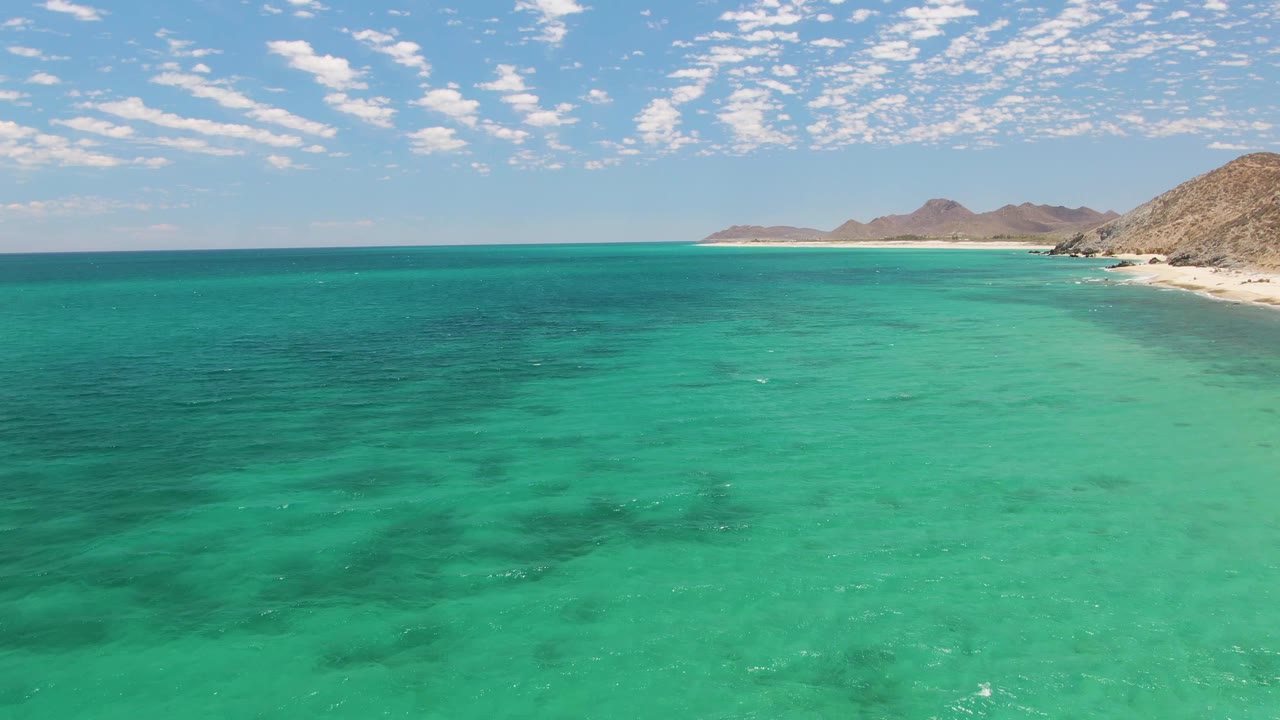 At one second, the Barakas Beach view holds on clear green-blue water, a pale beach strip along the right, distant rocky landforms, and no visible people.