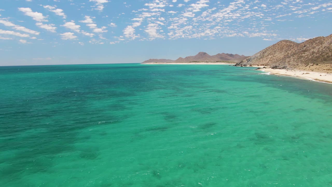 Aerial view of turquoise water off Barakas Beach in Baja California Sur, with sandy shoreline and rocky desert mountains in the distance and no visible people.