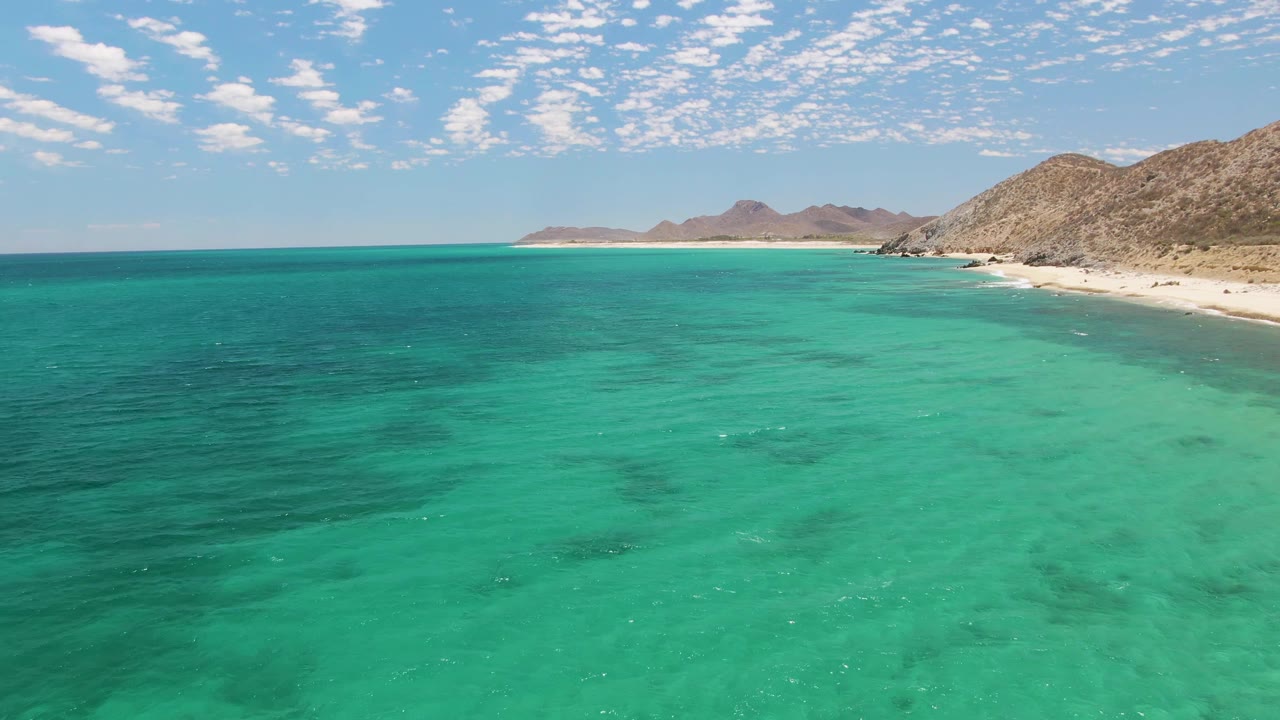 Wide aerial frame over green-blue water at Barakas Beach, with pale sand and low rocky hills along the right edge and no visible people.