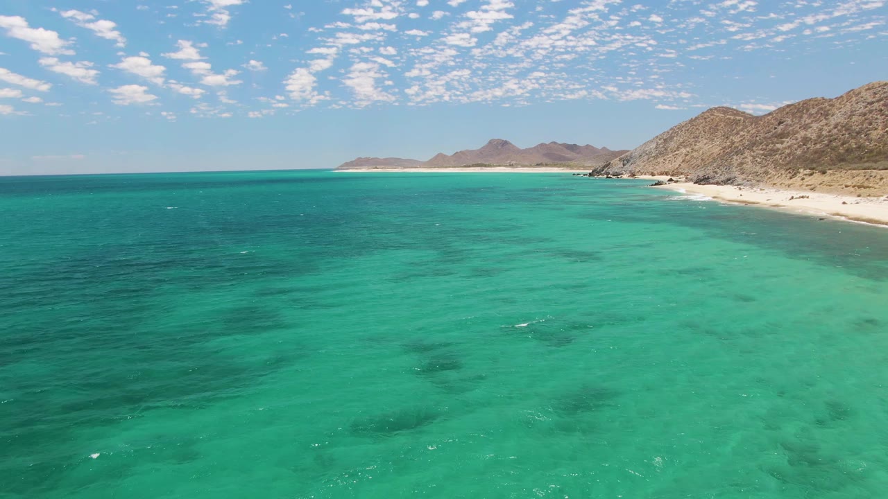 Aerial coastal view at Barakas Beach with shallow turquoise water, darker underwater patches, a narrow sandy beach, rocky mountains, and no visible people.