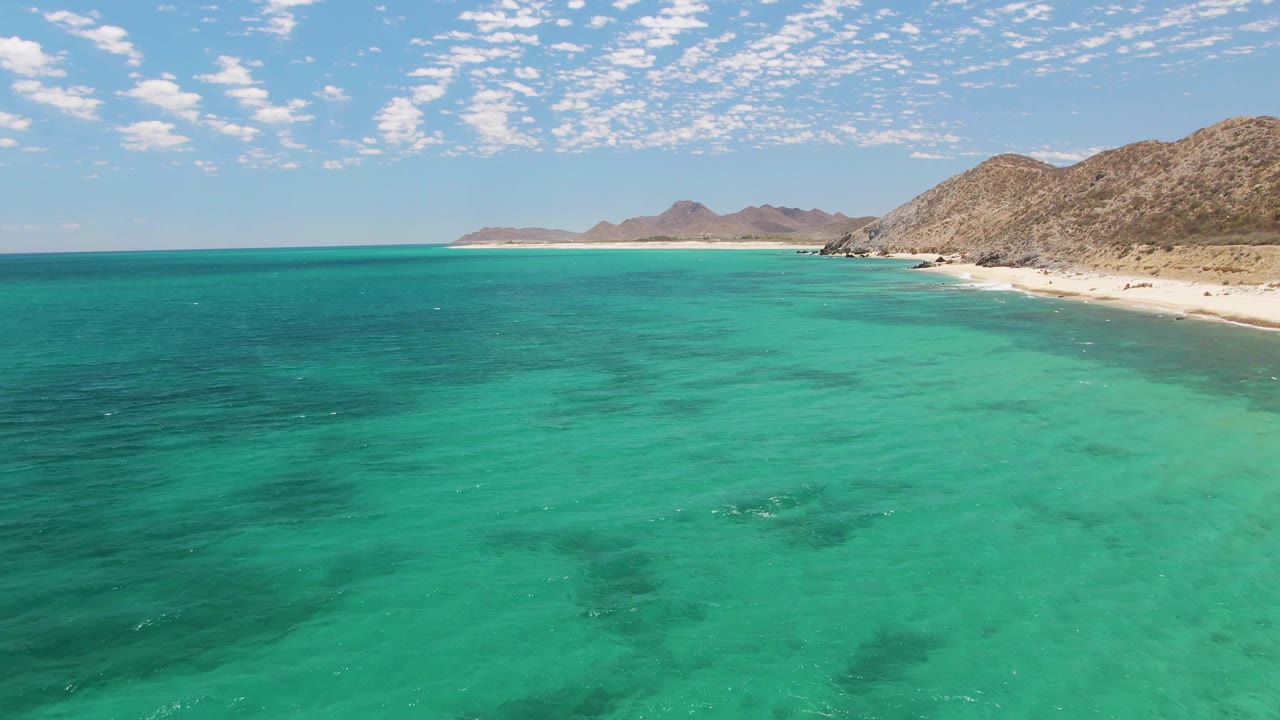 Wide drone view of Barakas Beach with rippled turquoise ocean in front, sandy shoreline on the right, distant headlands, rocky mountains, and no visible people.