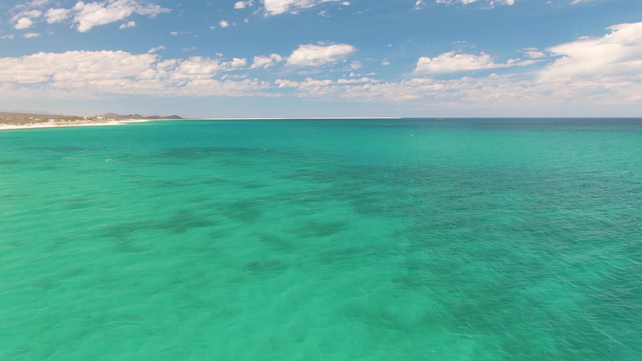 Turquoise water fills the opening frame at Barakas Beach, with a sandy shoreline far left, dark underwater patches, a level horizon, and no visible people.