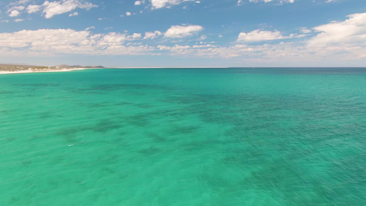 At 0:02, the frame shows a wide aerial view over turquoise Barakas Beach water, with darker submerged areas in the midground, coastline left, and no visible people.