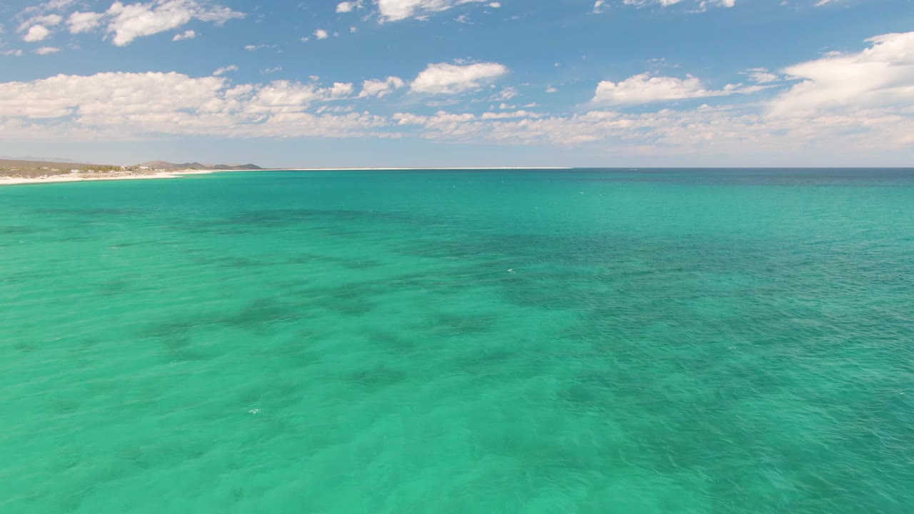 At 0:03, rippled turquoise water covers the foreground at Barakas Beach, with the sandy coast visible in the upper left and no visible people.
