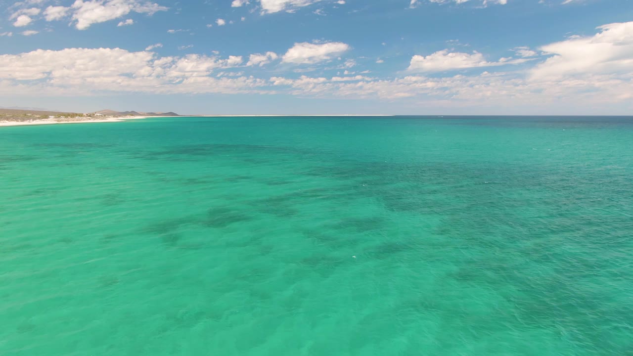 Opening frame over turquoise water at Barakas Beach, Baja California Sur, with darker underwater patches, a sandy shore on the left horizon, scattered clouds, and no visible people.