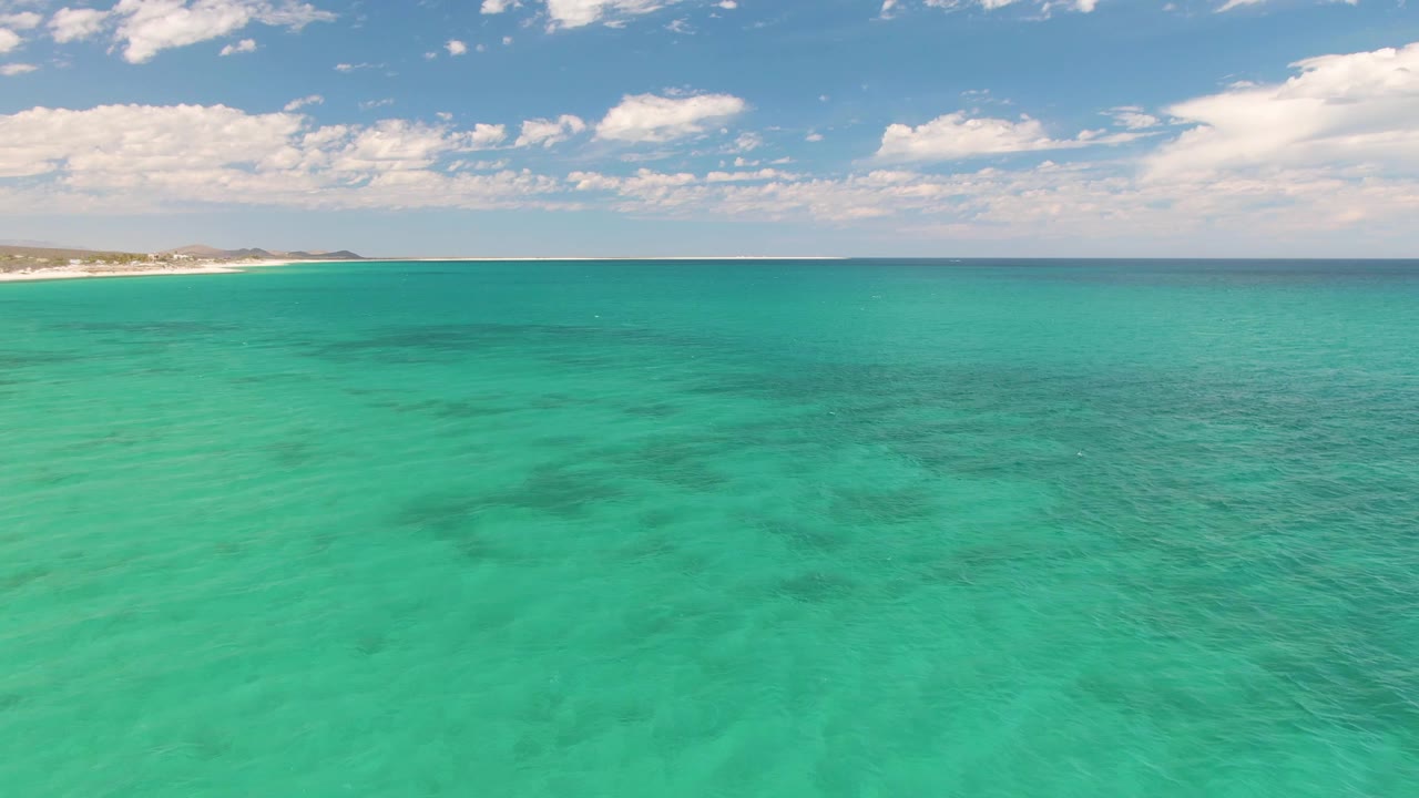 Clear turquoise sea beneath scattered clouds with beach in the distance.