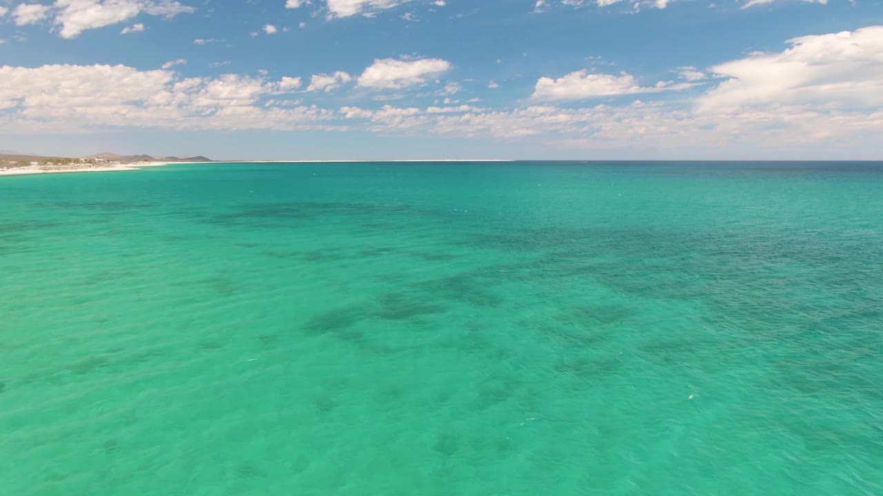At 0:02, turquoise ocean dominates the frame at Barakas Beach, Baja California Sur, with darker shapes under the water, a pale coast along the left distance, and no visible people.
