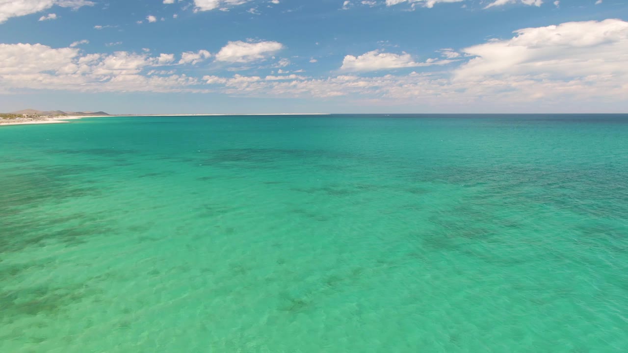 Aerial frame over clear green-blue water at Barakas Beach, Baja California Sur, with faint shoreline at left and no visible people.