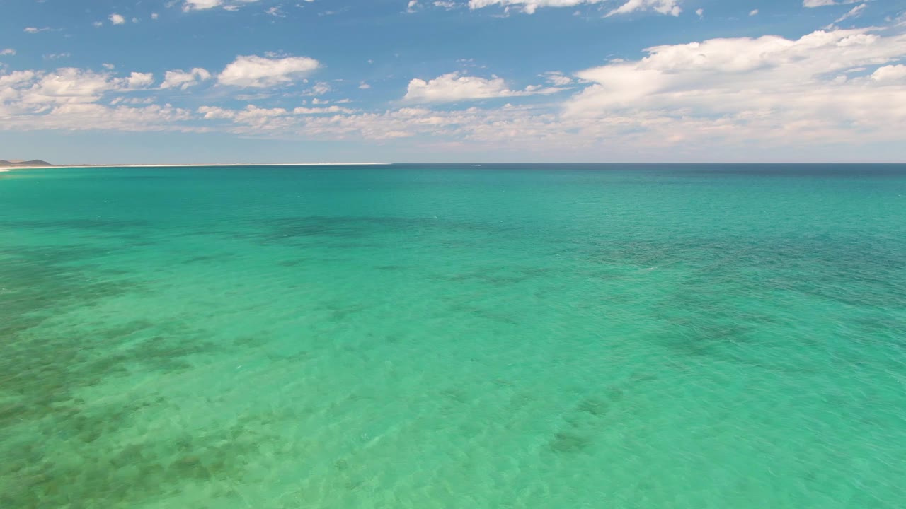 Mid-clip aerial view at Barakas Beach, Baja California Sur, with shallow turquoise water in the foreground, shore far left, and no visible people.