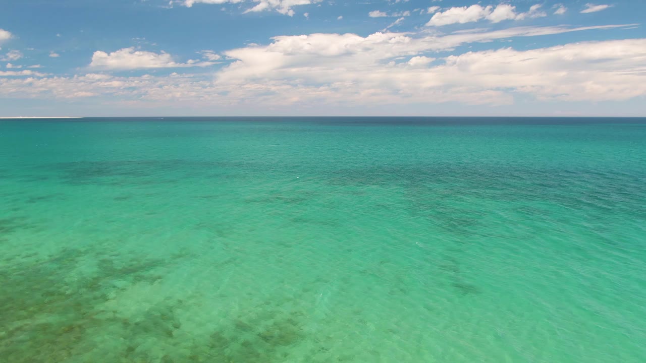 Opening aerial view of turquoise ocean at Barakas Beach, Baja California Sur, with darker underwater patches, a high horizon, scattered clouds, and no visible people.