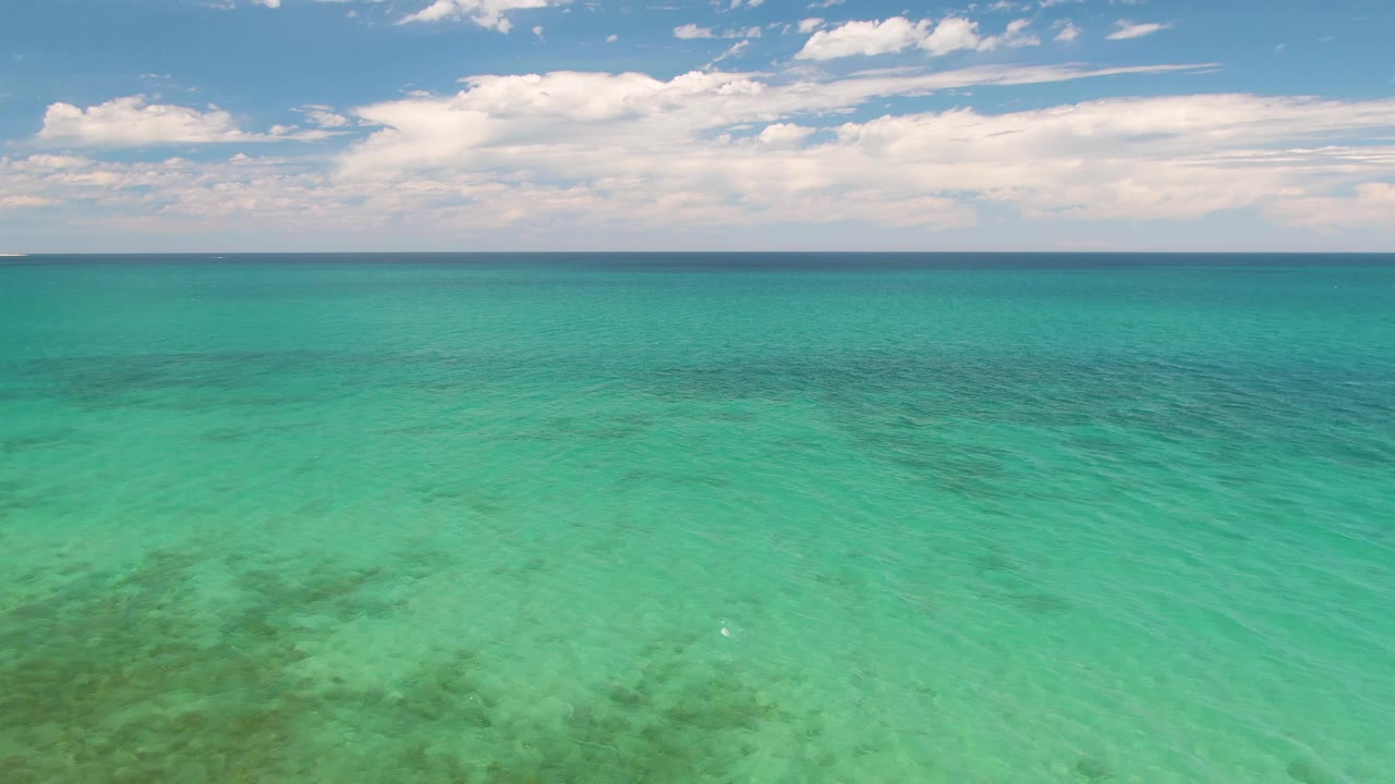Aerial frame of Barakas Beach water with broad turquoise color, soft ripples, a thin distant shoreline line, white clouds, and no visible people.