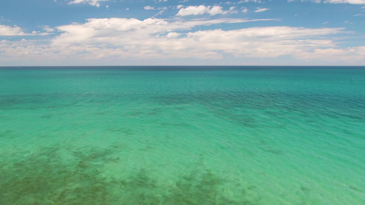 Mid-clip aerial view at Barakas Beach showing transparent green-blue water, darker seabed shapes, a level horizon, cloud bands, and no visible people.