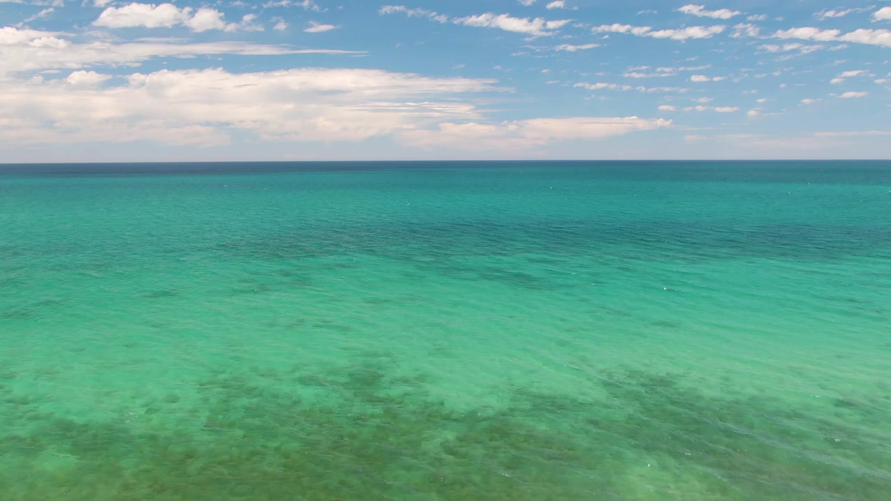 Opening frame of Barakas Beach shallow turquoise ocean with darker seabed patches under clear water, scattered clouds above the horizon, and no visible people.