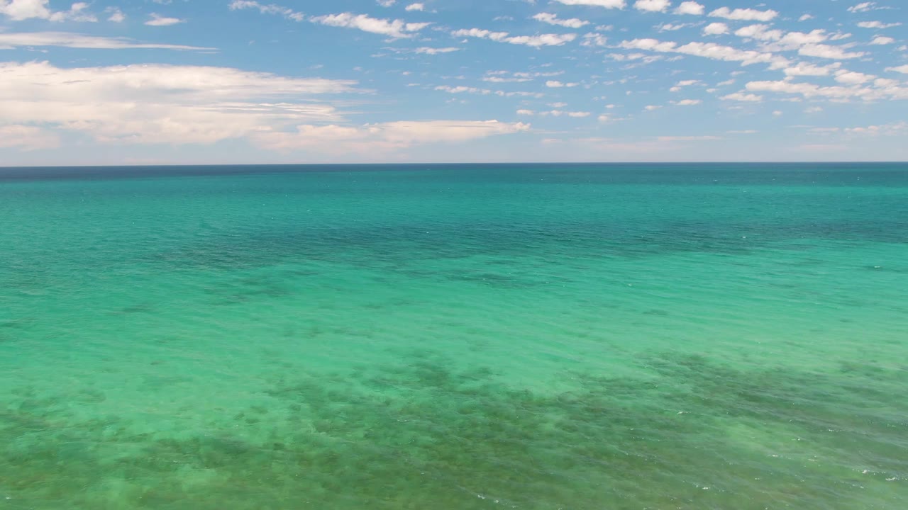 Wide view over Barakas Beach turquoise shallows, with darker underwater patches spread across the lower frame, small clouds in the sky, and no visible people.