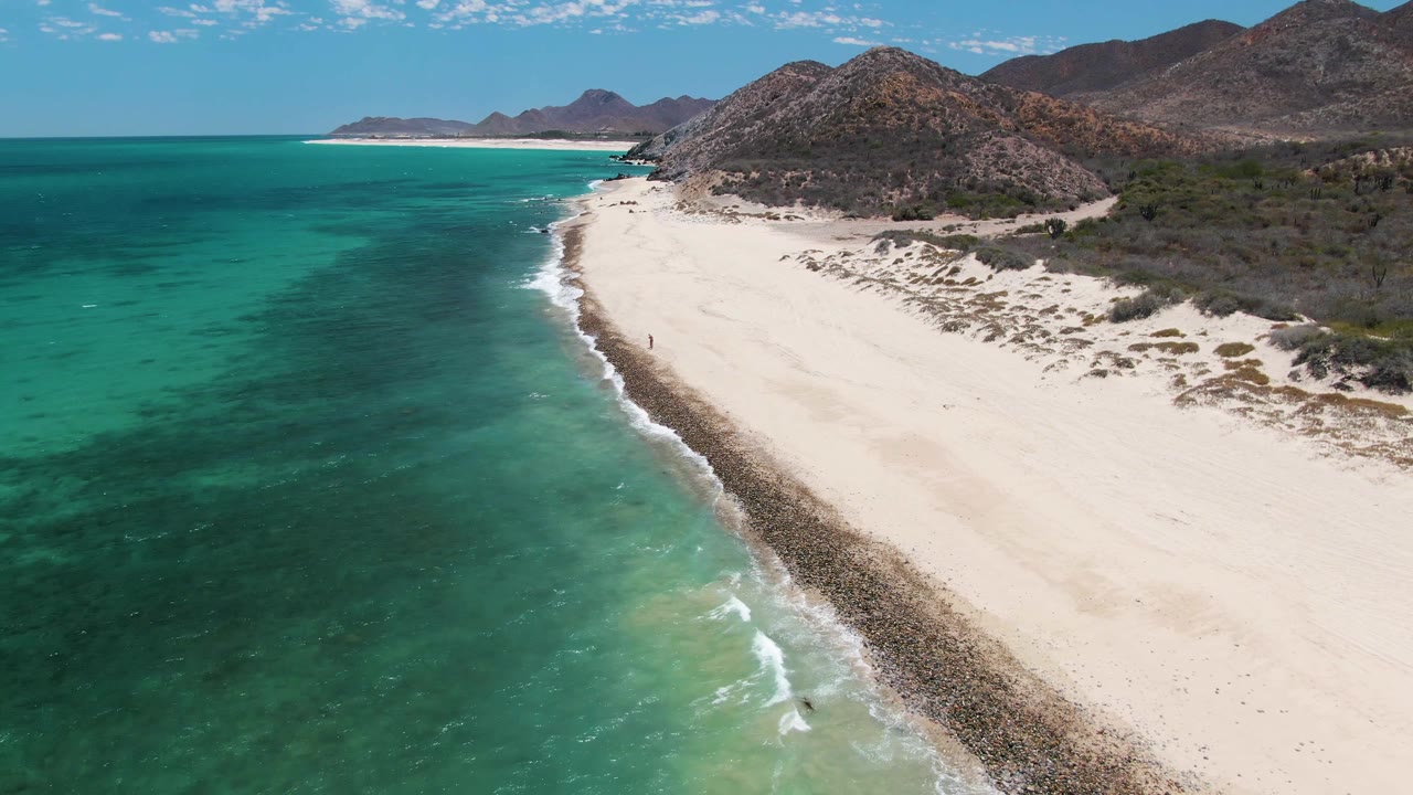 Aerial view at 0:01 over Barakas Beach, with darker turquoise water filling the left side, a thin white surf line along the sand, rocky hills behind the beach, and no visible people.