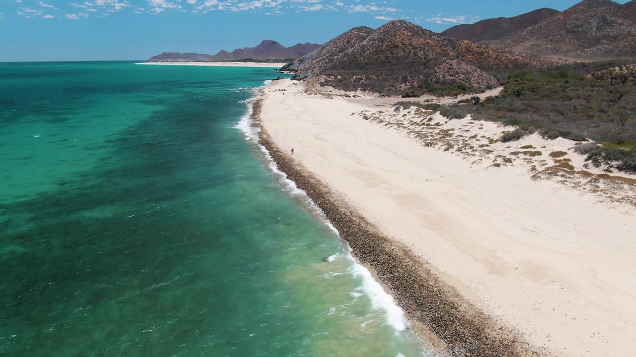 Mid-clip aerial frame over Barakas Beach, Baja California Sur, showing the coastline angled across the frame, turquoise water with subtle wave texture, bright sand, rocky shoreline, and no visible people.
