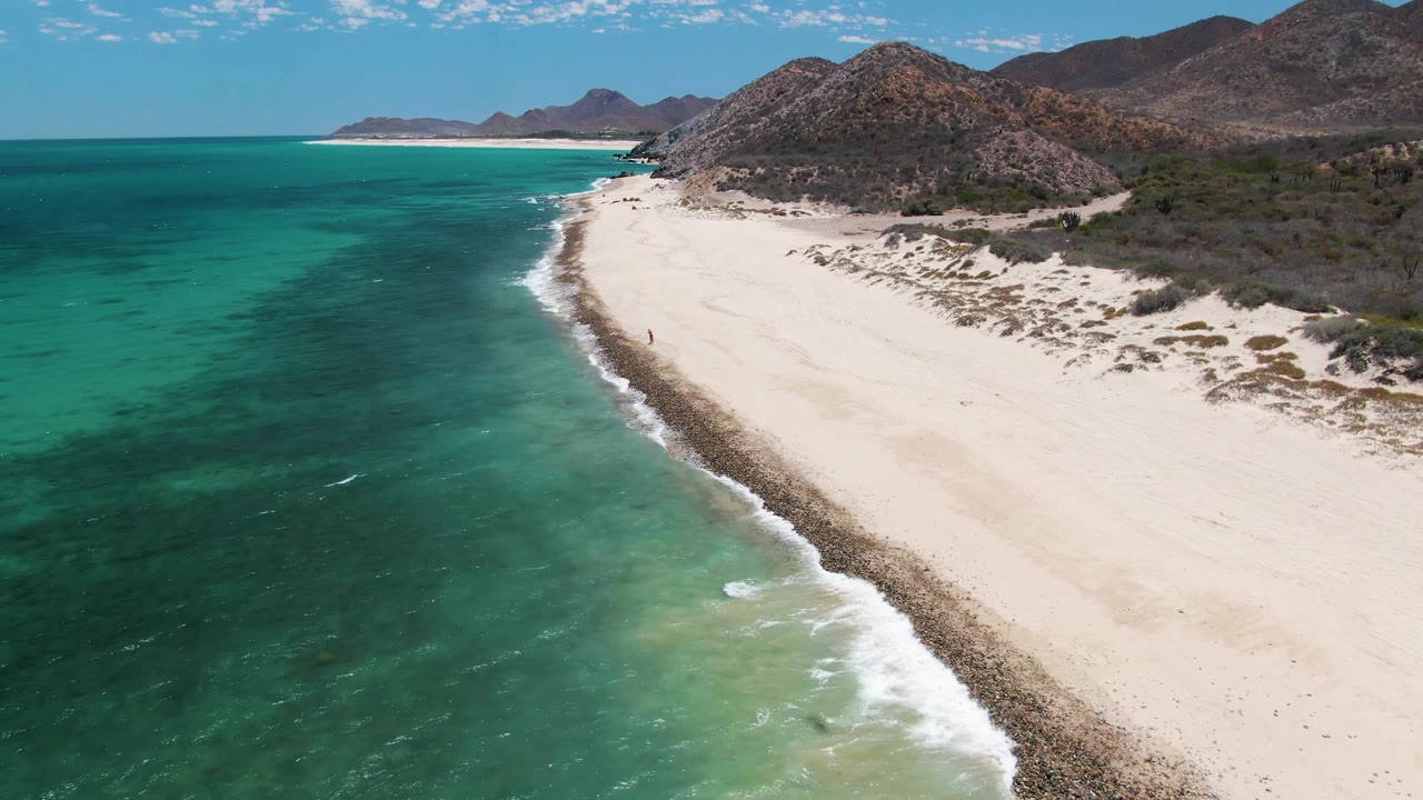 At 0:03, the aerial view shows green-blue water closer to the lower left, small white waves breaking along the rocky edge of Barakas Beach, mountains beyond the dunes, and no visible people.