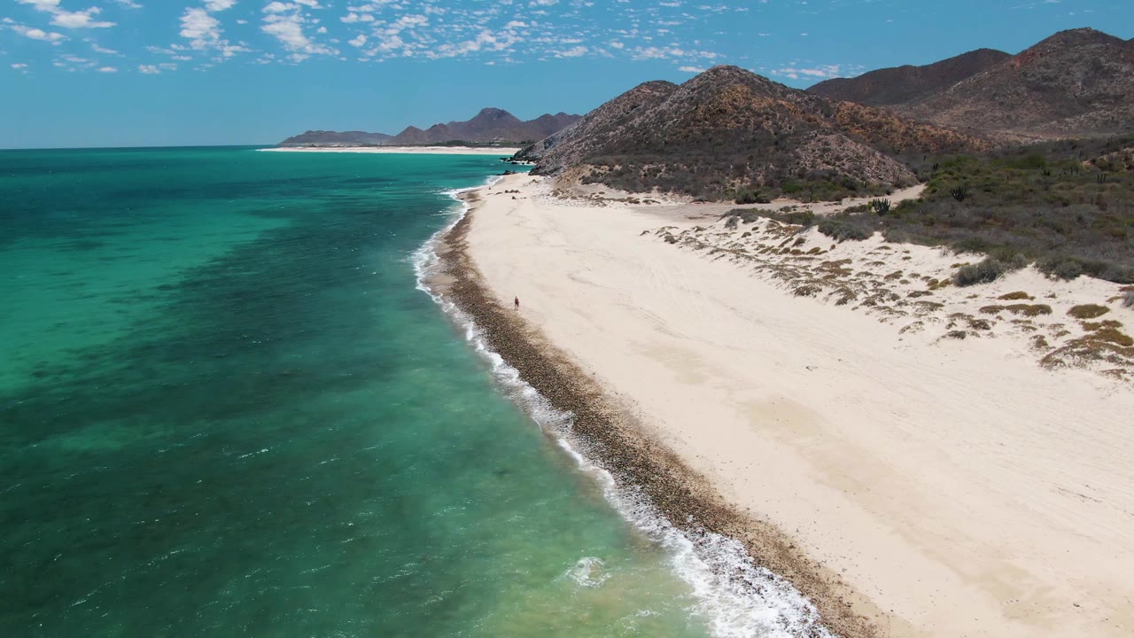 Aerial frame over Barakas Beach in Baja California Sur, with turquoise water at left, pale sand curving along a rocky surf line, hills in the background, and a small person on the beach.