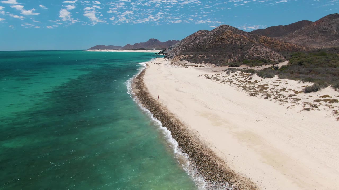 A wide Barakas Beach aerial frame with open turquoise sea to the left, a pale beach sweeping toward distant mountains, dry rocky hills to the right, and one small walker on the sand.