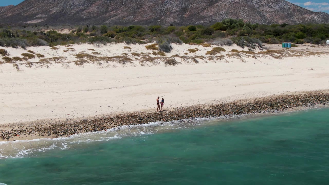 At Barakas Beach in Baja California Sur, two people stand near the rocky edge of the shore as turquoise water fills the foreground and low dunes lead back to mountains.