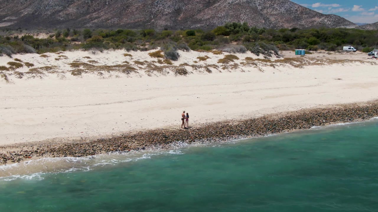 Two people on the shoreline below dunes, vehicles, vegetation, and mountains.