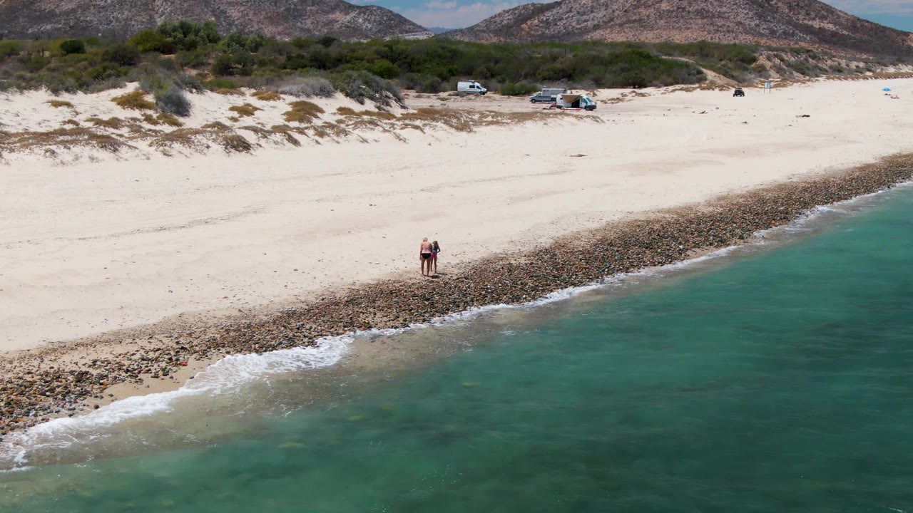 Two people are near the rocky edge of Barakas Beach as turquoise water reaches the sand and vehicles sit near the dunes in the distance.
