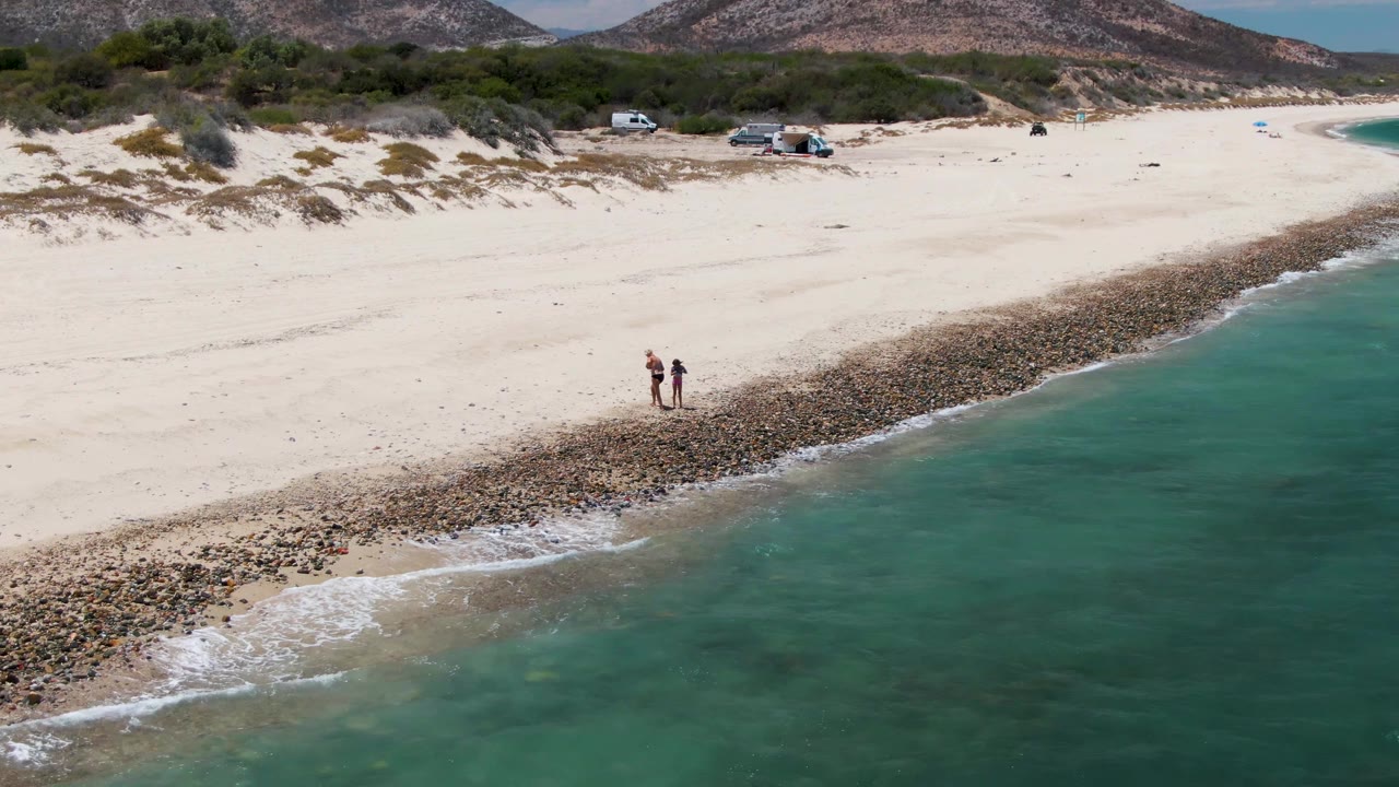 Two people on pale sand beside a rocky strip and clear ocean water.