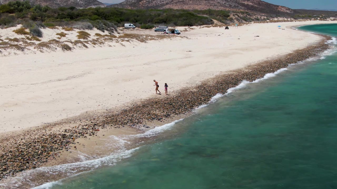 The walkers continue along Barakas Beach beside the pebble-like shoreline strip while a small wave breaks along the turquoise waterline.