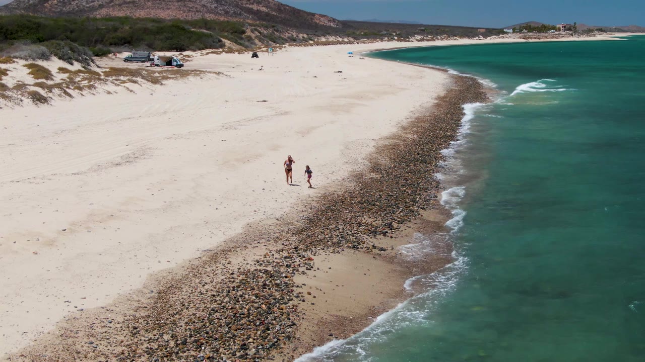 Aerial view at Barakas Beach, Baja California Sur, with two people walking on pale sand beside a curved pebble shoreline and turquoise water.