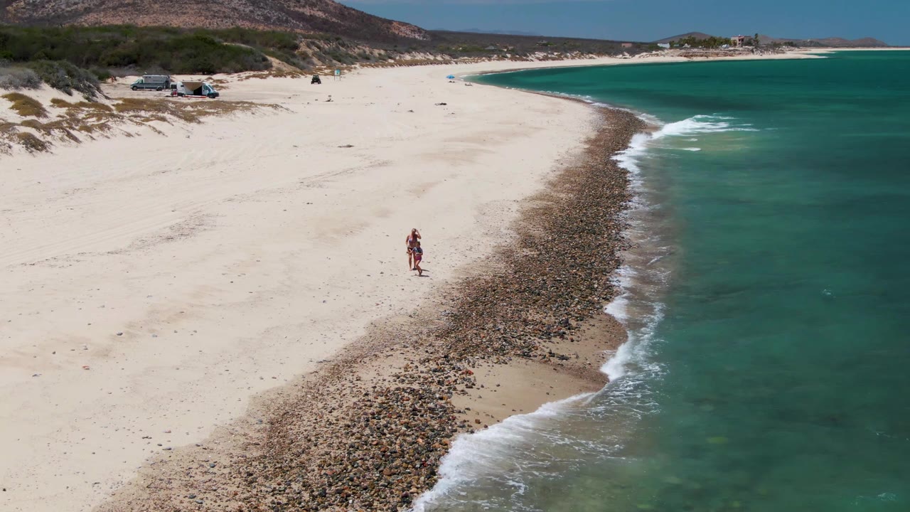Two people near the pebble edge of a sandy beach with waves rolling in.
