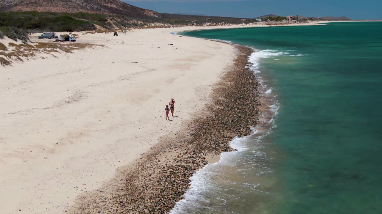 Aerial view over Barakas Beach with two people farther along the curving sand, the pebble band widening between the beach and turquoise surf.