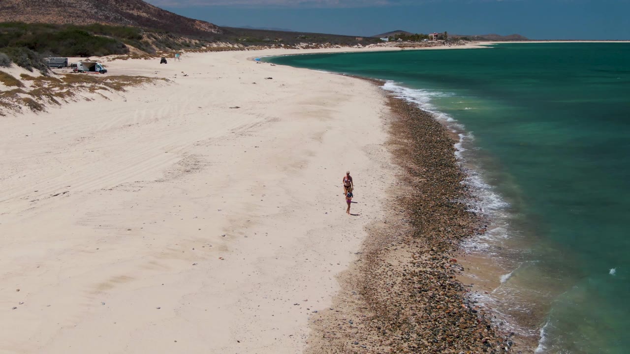At Barakas Beach in Baja California Sur, two small figures walk along pale sand beside a rocky waterline with turquoise ocean to the right and low hills in the distance.