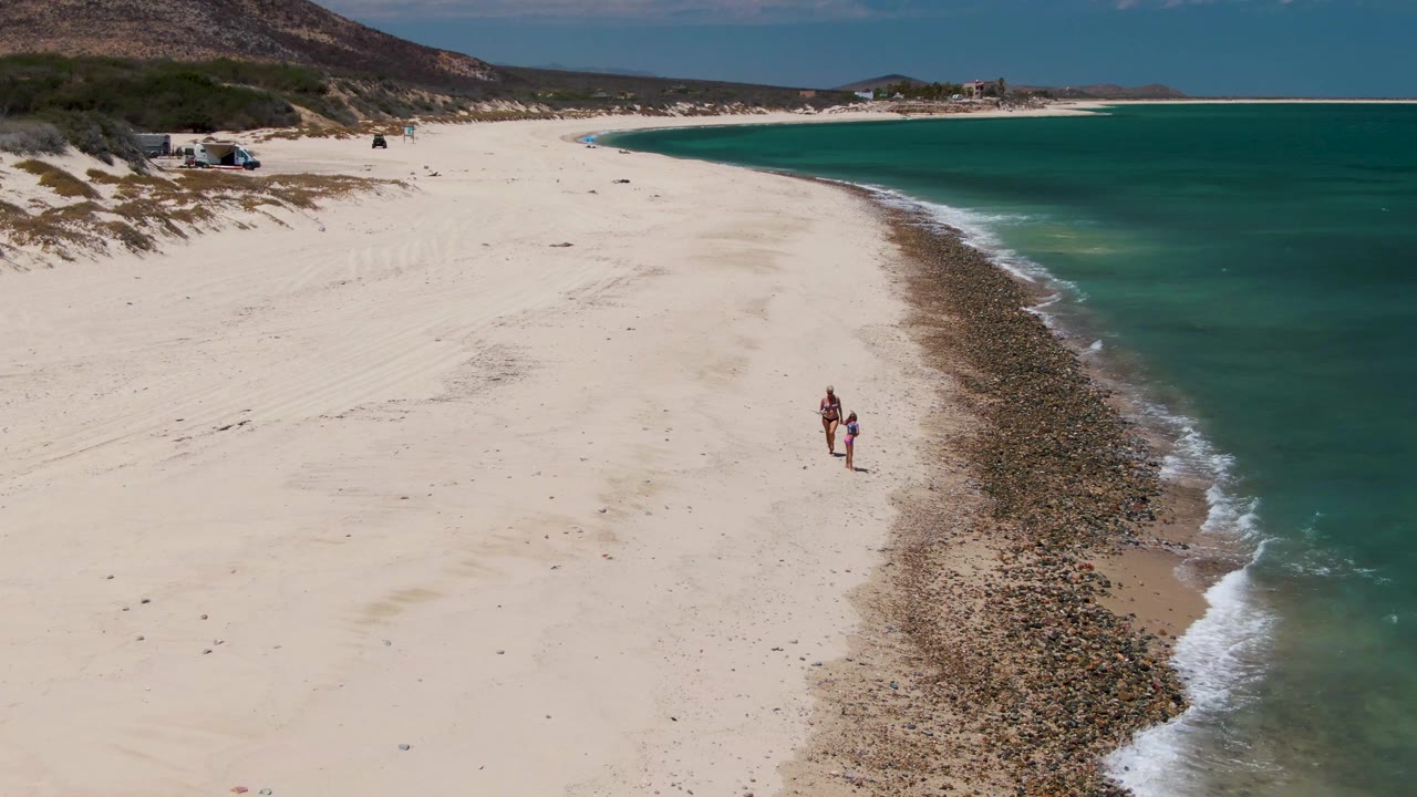 Two people appear slightly farther along the Barakas Beach shoreline, standing out against the pale sand while turquoise water curves along the right side of the frame.