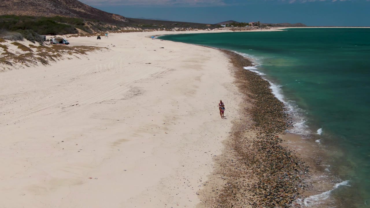 Aerial frame at Barakas Beach, Baja California Sur shows one person walking on pale sand beside a dark rocky shoreline with turquoise water on the right and dunes in the distance.