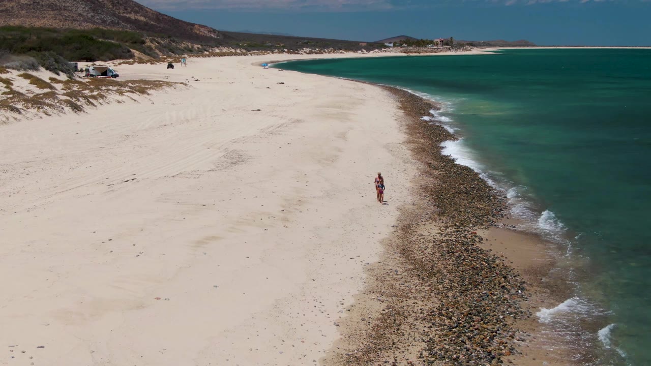 Person walking along pale sand beside rocks and turquoise ocean.