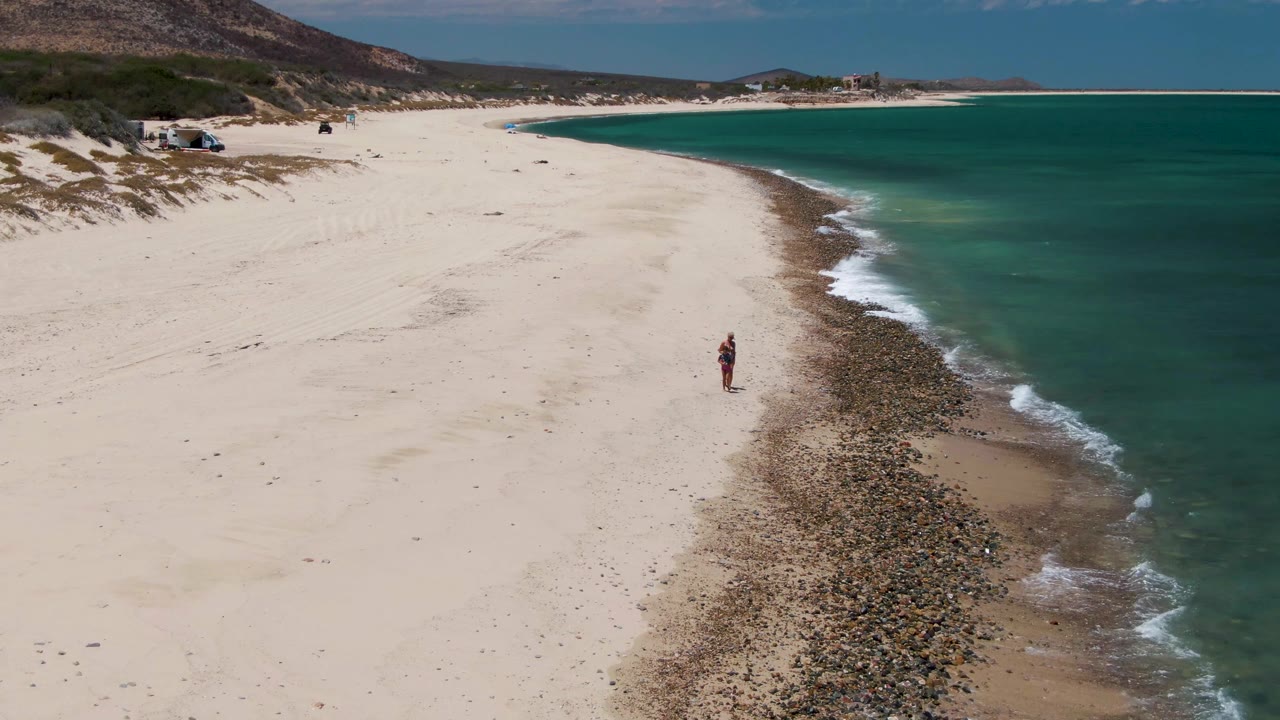At 0:02, the aerial composition keeps the Barakas Beach walker near the rocky tide edge while the green-blue ocean fills the right side and low hills line the background.