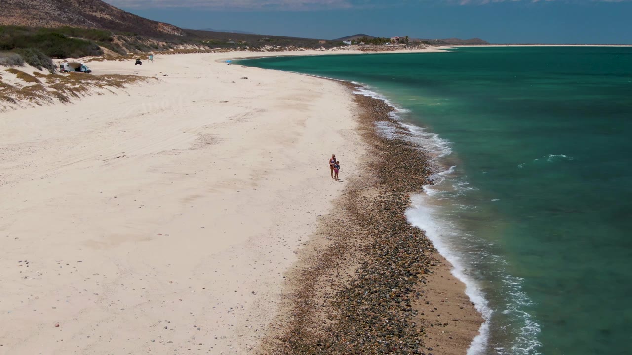 Aerial frame of Barakas Beach in Baja California Sur with a lone person on white sand near a rocky shoreline strip and turquoise ocean to the right.