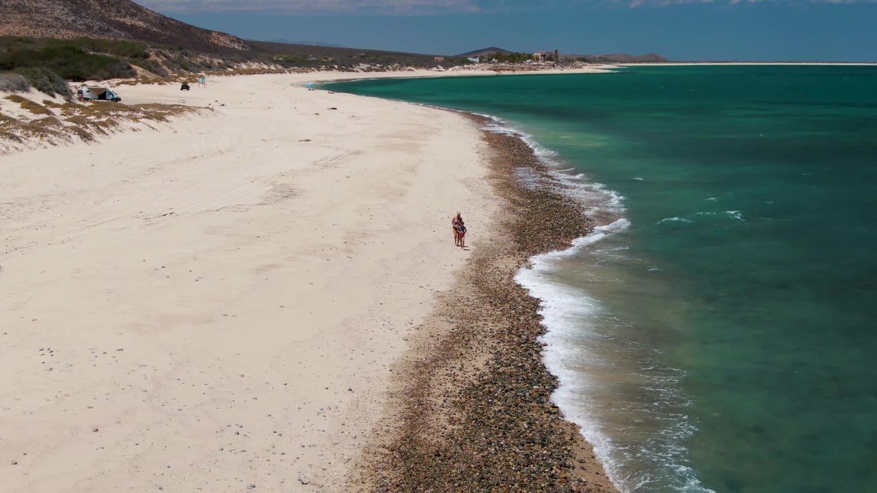 Drone view of a person walking beside waves on a pale sandy beach.