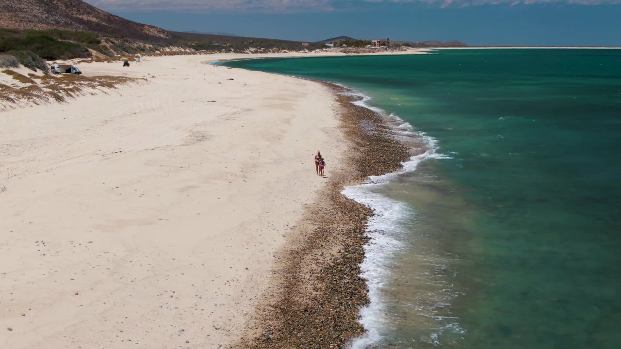 Elevated view of the Barakas Beach shoreline as the walker remains near the center of the beach, with a curved rocky band and green-blue water filling the right half of frame.