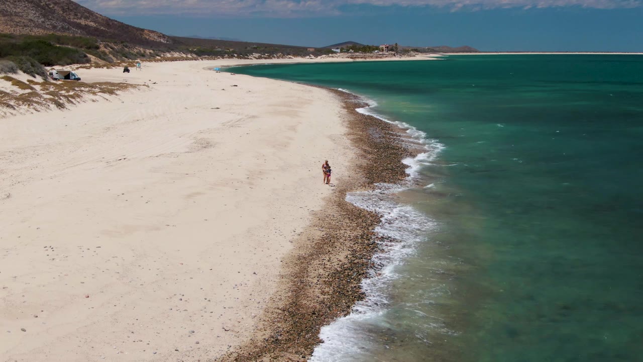 Wide aerial beach frame with a person walking near the surf and hills inland.